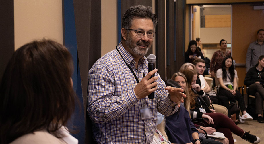 A man in a plaid shirt holds a microphone in a crowded room. 
