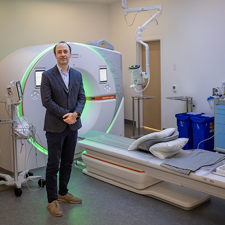 Tall man with dark hair standing hands clasped in front of large CT scanner.