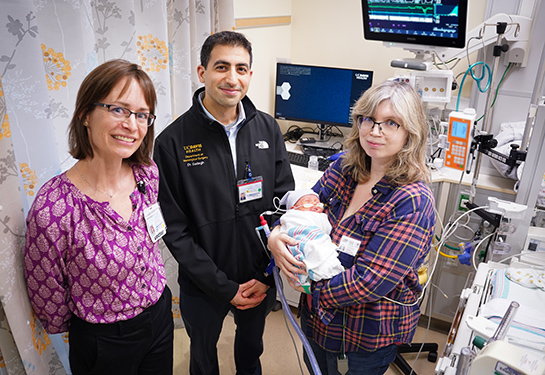 Female and male doctors in casual clothes stand next to a mother holding her infant baby in a hospital room.