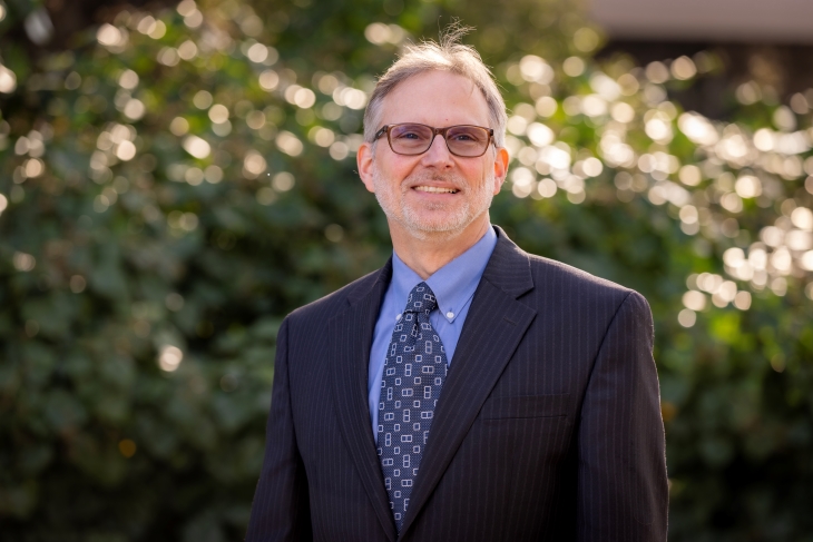 A man in a dark suit, blue shirt and tie stands outdoors in front of sunlit green foliage.