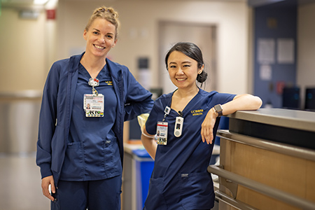 Blonde woman and black haired woman both in blue scrubs pose in a hospital hallway.