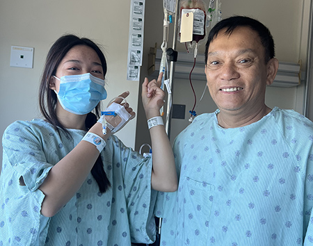 Young Asian woman in a surgical mask and hospital gown stands next to an Asian man in a hospital gown after they both had a procedure in the hospital.