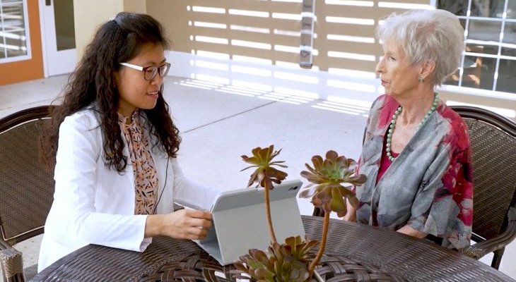 A healthcare professional sits outdoors at a round patio table, showing information on a tablet to an older adult.