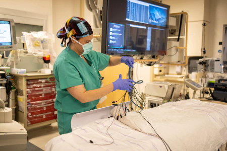 A clinician in scrubs prepares monitoring wires beside a patient bed in an electrophysiology lab.