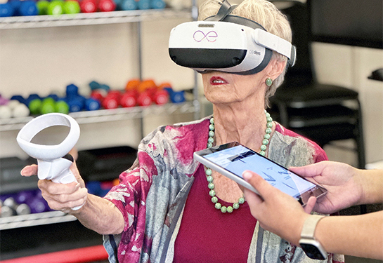 A senior woman wearing a virtual-reality headset raises a controller in her right hand.