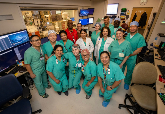 A large team of hospital staff in scrubs gathered together in a control room of an electrophysiology lab.