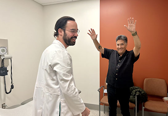 A physician stands in front of a patient in a clinic, while the patient holds his arms in the air to show how high he can reach. 