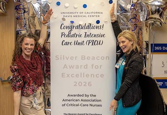 Two nurses stand next to pull up banner that states “Congratulations, Pediatric Intensive Care Unit!”