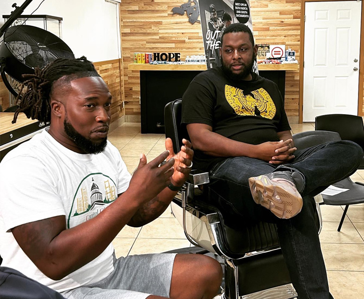 Two men talk in a barbershop, one gesturing while the other listens from a barber chair.