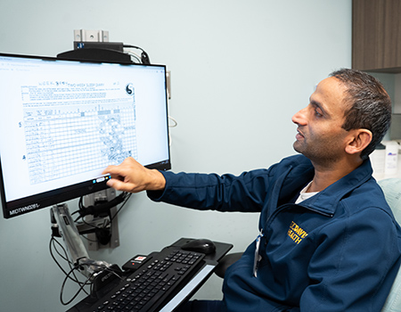 A man wearing a navy blue jacket with UC Davis Health brand in yellow. He is pointing to a screen showing a table