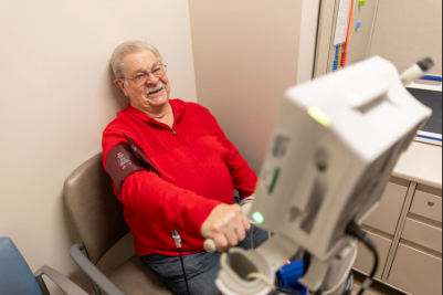 An older adult sits in a clinic chair with an arm extended into a blood pressure cuff machine while medical equipment and cabinetry are visible in the exam room.