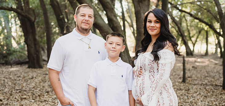 A father, expecting mother and 10-year old son are standing with trees behind them. Father and son in white polos and blue jeans. Mother in a dress with her hands around her belly.