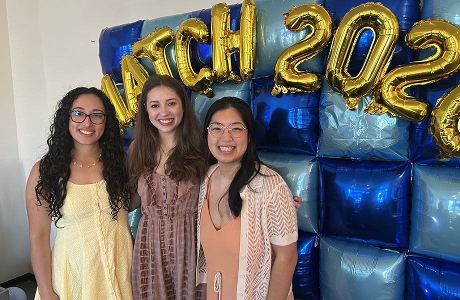 Three women stand together in front of a blue balloon wall with gold balloons that spell out Match Day 2026