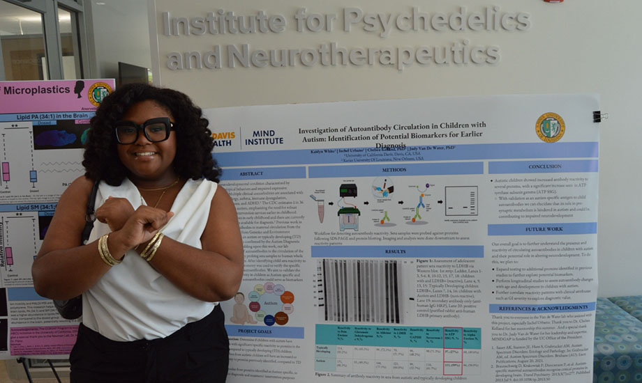 Kaidyn White, a MINDGAP scholar, stands next to a research poster she created, while crossing her arms over her chest to create a letter X