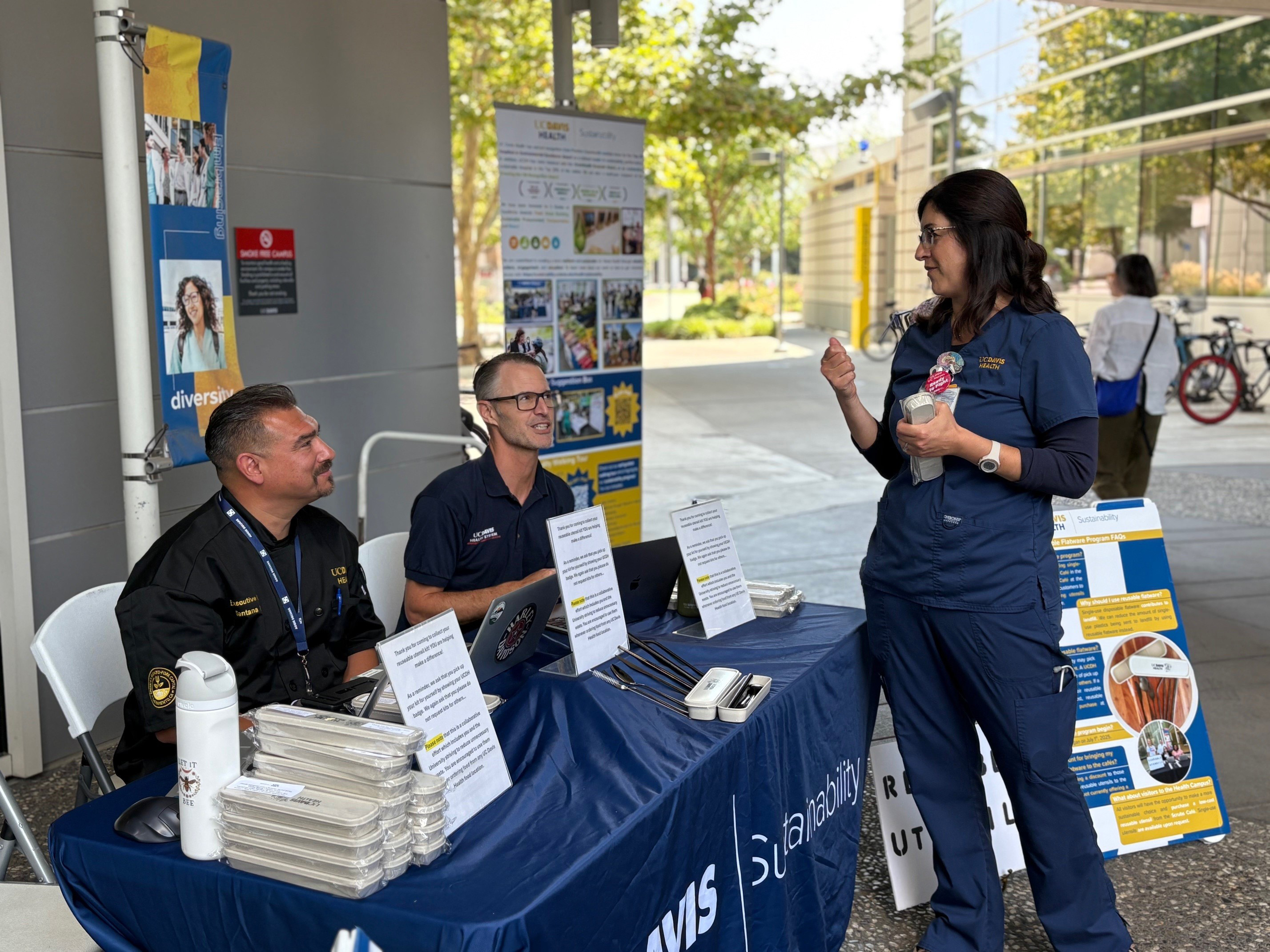 Two adults sit behind a table containing reusable utensil kits while another adult talks with them. 