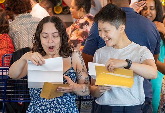 Two people react with excitement while opening envelopes at the on-campus Match Day event in a large room of an apartment building