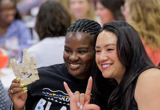Two women smiling at an event, one holding a small wooden block structure.