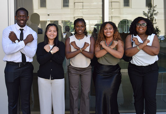 A group of five college students stands outside next to a glass building, crossing their arms over their chests to make the letter X