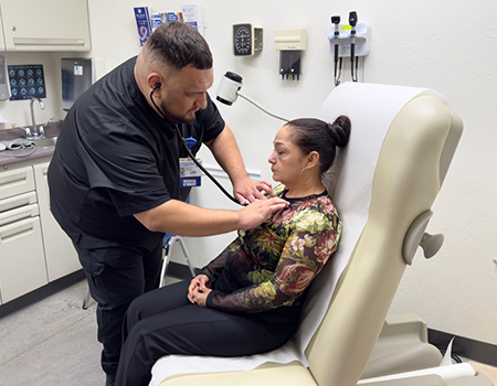 A medical student in black scrubs leans into a medical exam chair to check the heartbeat of a woman who is sitting down.
