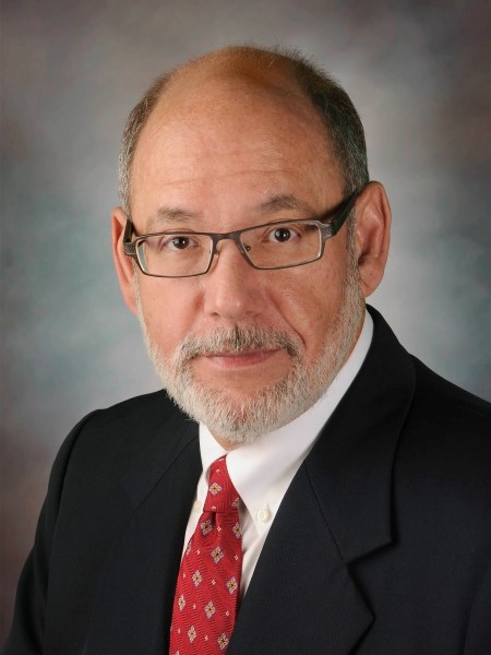 Studio portrait of an individual in a dark suit and red tie, wearing glasses, against a neutral background.