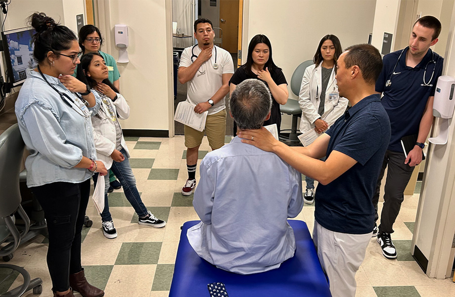 Seven medical students with stethoscopes surround two faculty doctors teaching how to perform neck examinations.