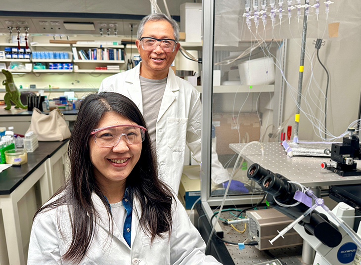 Two people in lab coats wearing safety glasses stand beside laboratory equipment and a microscope in a research lab.