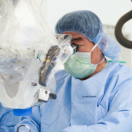 Dr. Kee Kim is in the operating room, looking through a microscope. He is wearing blue scrubs, a mask and a pair of gloves.