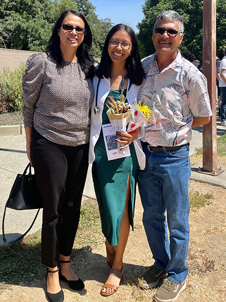 Medical student Abigail Vidrio, wearing a white coat and stethoscope at her induction ceremony, surrounded by her parents. 