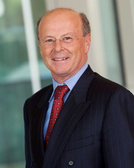 Professional headshot of individual in dark suit and red tie, standing indoors against a softly blurred background.