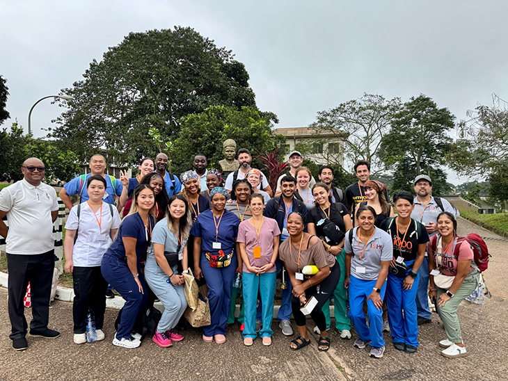 A large group of healthcare professionals wearing scrubs and badges pose outdoors, with trees and buildings in the background.