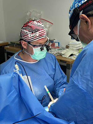 Two medical professionals in blue surgical attire perform a procedure under operating room lights using precision instruments.