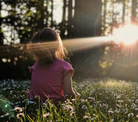 A young girl sitting on the grass, surrounded by trees with the sun beaming bright through the leaves