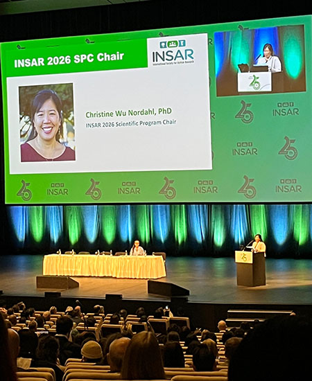 Christine Wu Nordahl speaks behind a podium on stage at the INSAR conference.  A large screen above projects her image, name and title and INSAR 2026. 