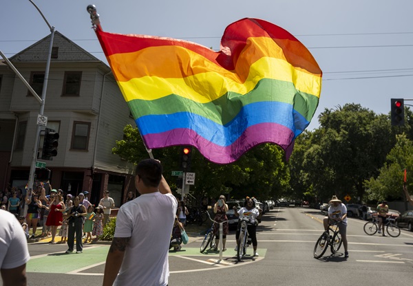 Large rainbow Pride flag is waved above a street as people walk and ride bicycles nearby.