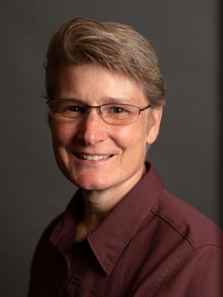 Studio portrait of an individual wearing glasses and a maroon button-down shirt against a dark background.