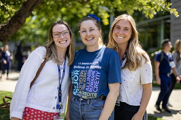 Three employees stand together outdoors wearing UC Davis Health apparel, posing for a photo on campus.