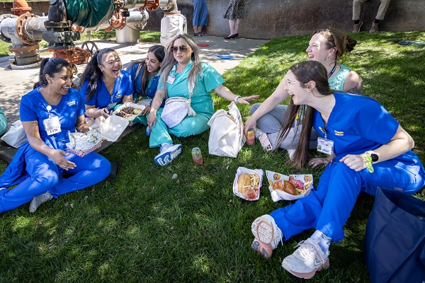 A group of healthcare employees sit on grass outdoors, eating food and talking together near campus buildings.