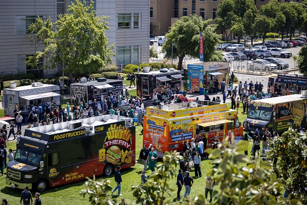 A wide aerial view shows multiple food trucks lined up on campus lawns with large crowds gathered around them.