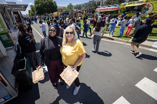 Two employees stand on a street near food trucks, holding boxed meals as crowds line up behind them.
