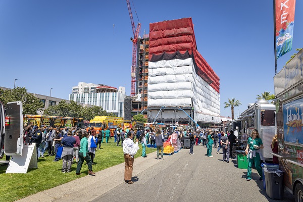 A large outdoor crowd stands near food trucks with a construction-wrapped building and crane in the background.