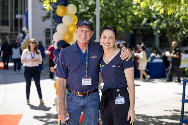 Two employees stand with arms around each other outdoors near event decorations and tables on campus.