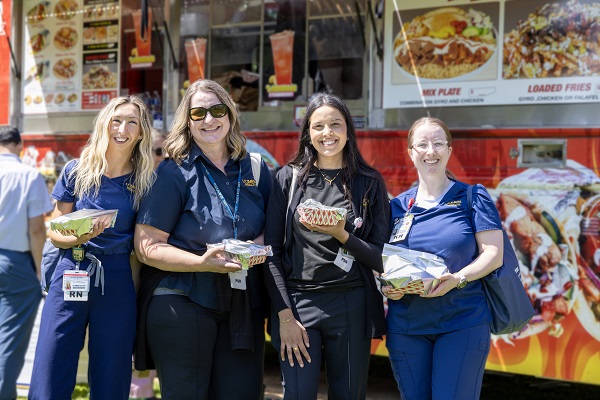 Four employees stand in front of a food truck holding boxed meals and smiling toward the camera.