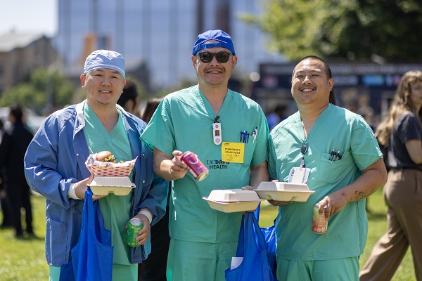 Three healthcare employees in scrubs stand outdoors holding food containers and blue tote bags.