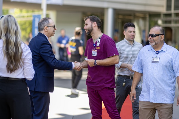 Two people shake hands on a red carpet walkway as others stand nearby at an outdoor campus event.