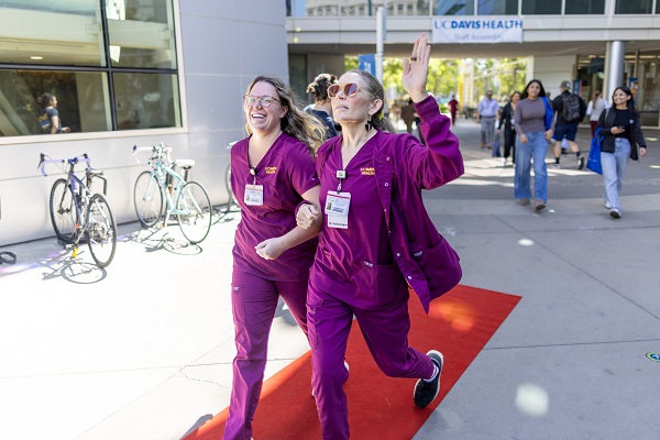 Two healthcare workers in scrubs walk along a red carpet outside the UC Davis Health main entrance as others follow behind.