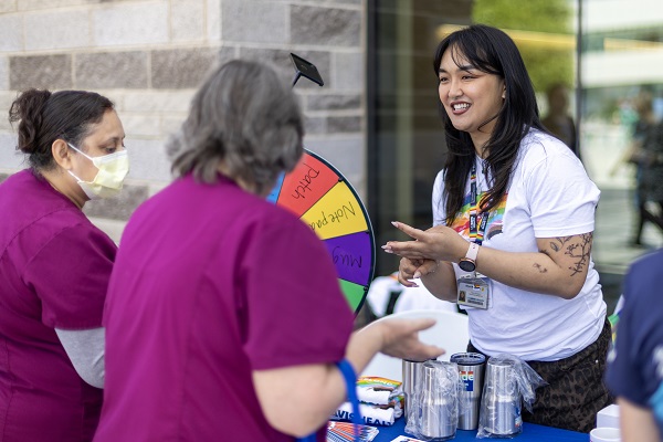 An employee speaks with visitors at a table while gesturing toward a colorful prize wheel during an outdoor event.