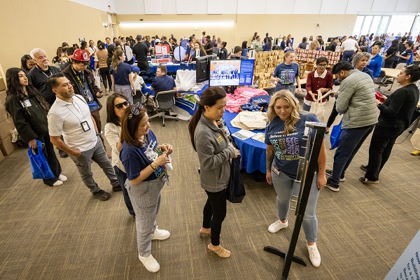 A large indoor event space filled with employees walking between information tables and displays.