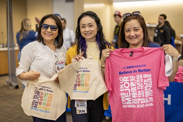 Three employees hold UC Davis Health &ldquo;Believe in Better&rdquo; tote bags and shirts at an indoor event.