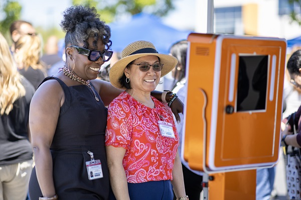 Two attendees pose together at an orange photo booth during an outdoor event.
