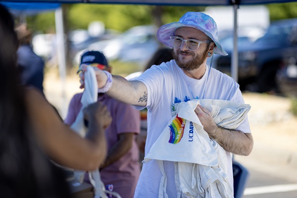 An attendee hands out white UC Davis Health tote bags with a rainbow graphic at an event booth.